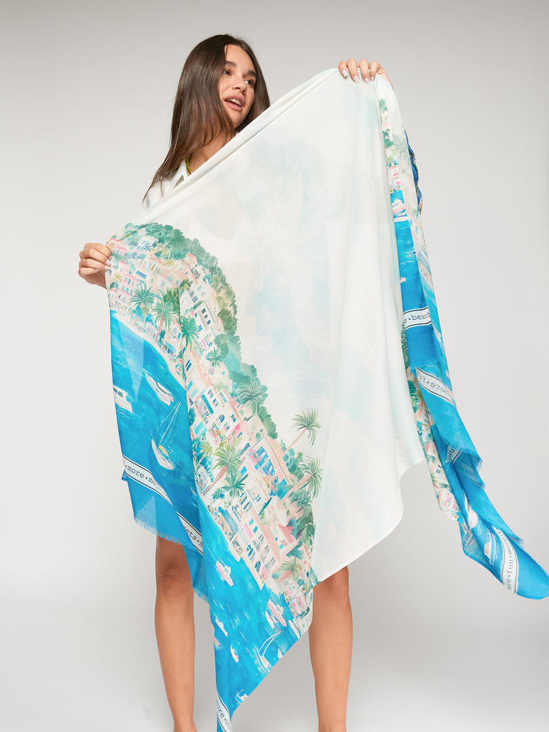 Woman holding Vilagallo Puerto Banus oversized cotton-modal scarf featuring an illustrated Mediterranean coastal village overlooking the sea in vibrant blue, green, yellow, and coral tones.