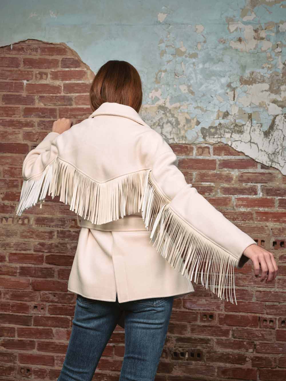 Back view of a model wearing the BLANK NYC Rodeo Star Blazer in Off White, showing long fringe against a brick wall.