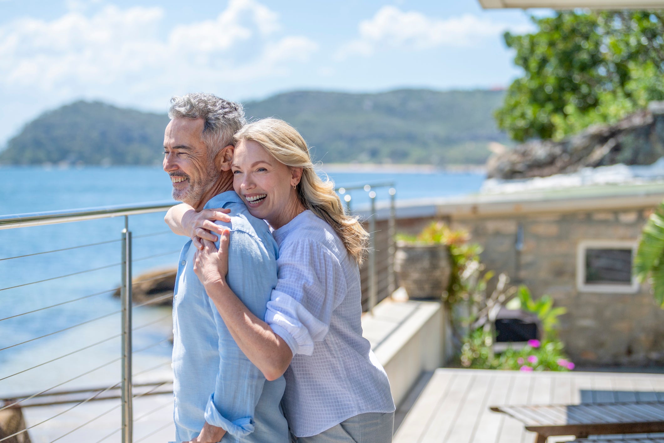 A smiling couple embracing on a wooden deck overlooking a scenic water view with mountains in the background. The man wears a light blue button-up shirt, and the woman wears a lightweight pale lavender-blue long-sleeve top.