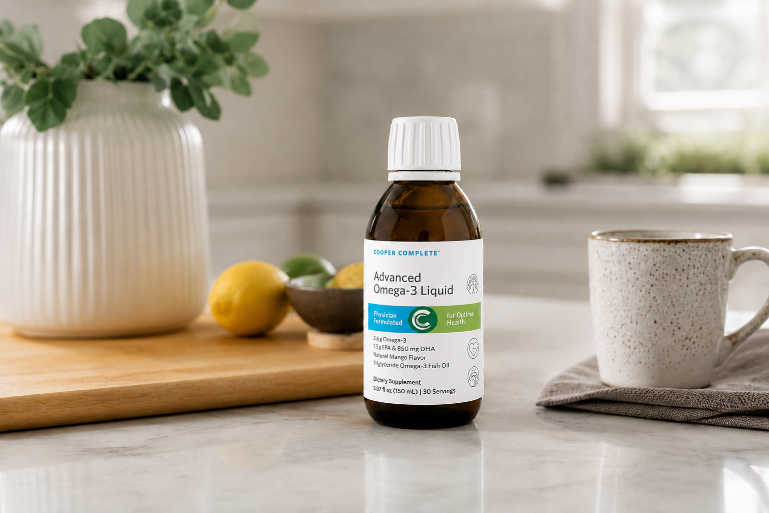 A bottle of Cooper Complete Advanced Omega-3 Liquid supplement on a kitchen counter, with a white ceramic vase of greenery, lemons and limes, and a speckled mug in a softly blurred kitchen background.
