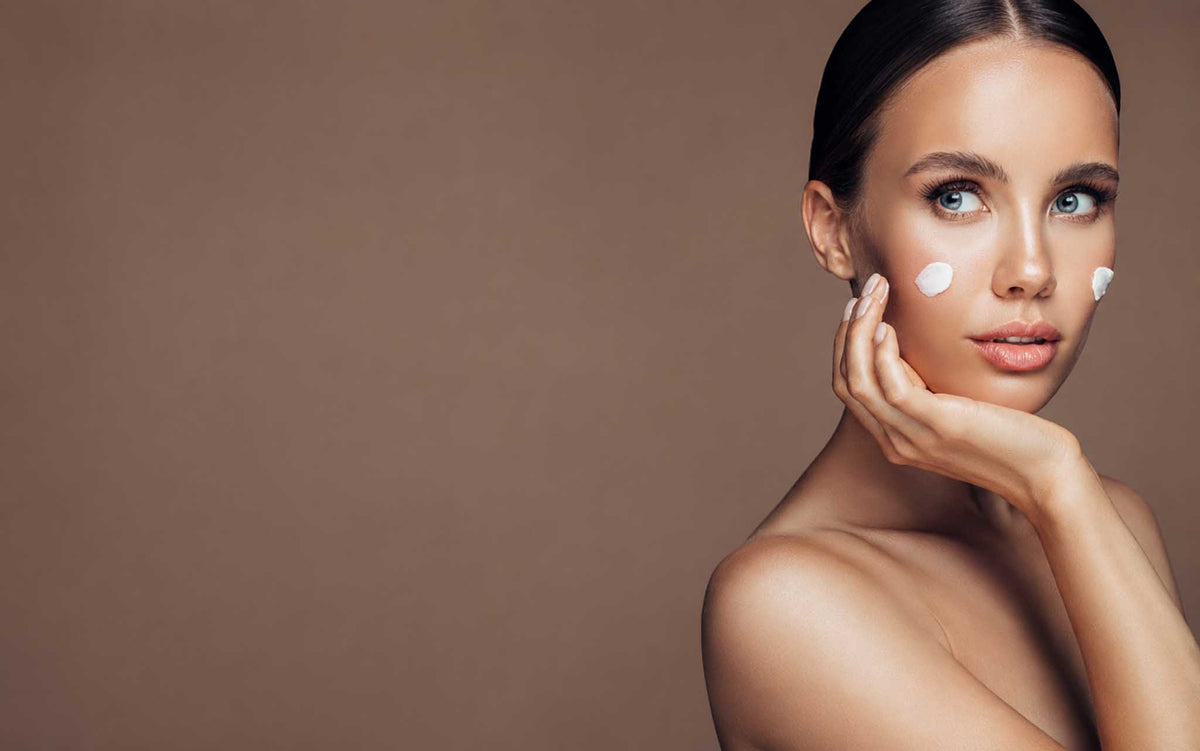 Model with slicked back dark hair applies skincare to healthy-looking skin against a mocha brown colored background.