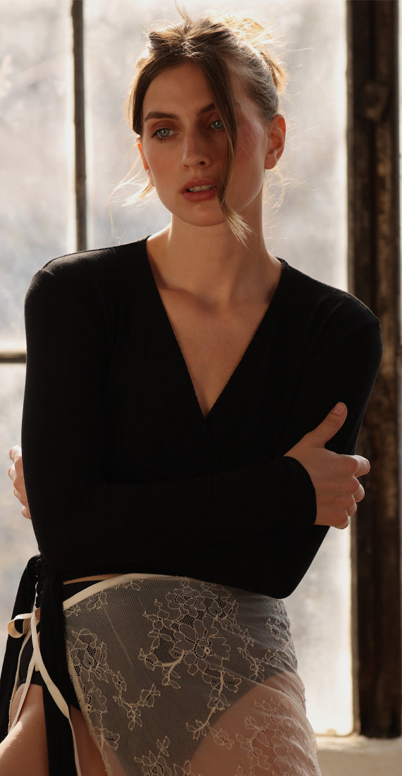 Front view of model wearing the black onyx Be Fit x Glee Ballerina Wrap with a deep V-neckline and waist tie, styled with a sheer lace skirt.