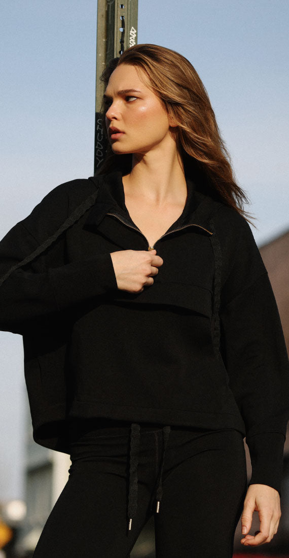 Model wearing the Be Fit x Glee black Onyx Raincoat outdoors with the front neckline a quarter unzipped.