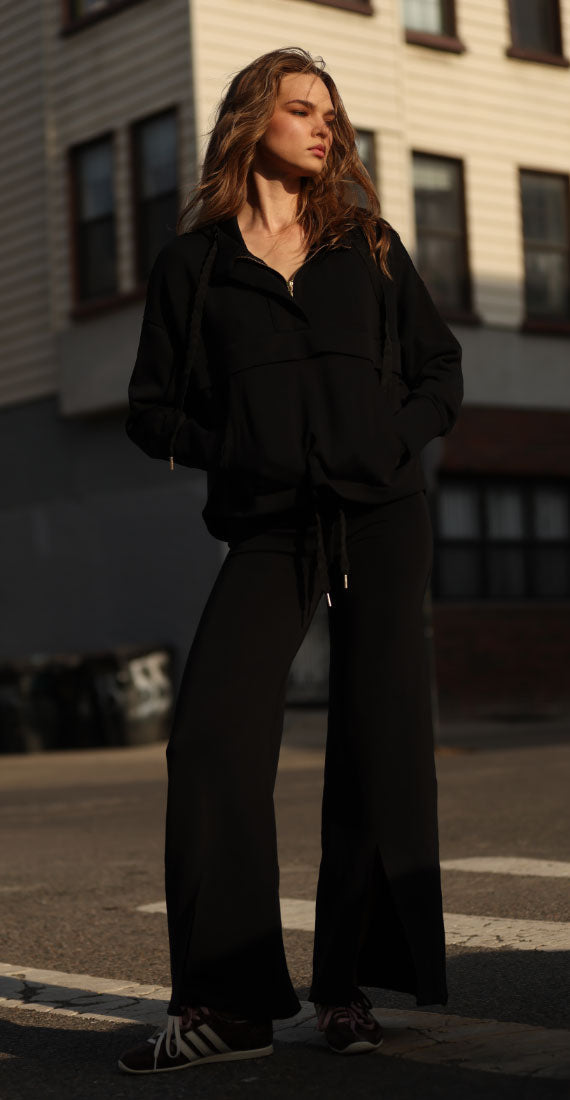 Front view of model wearing black onyx Be Fit x Glee Leggings Flare Soft Motion with a matching black onyx hooded jacket and sneakers outdoors.