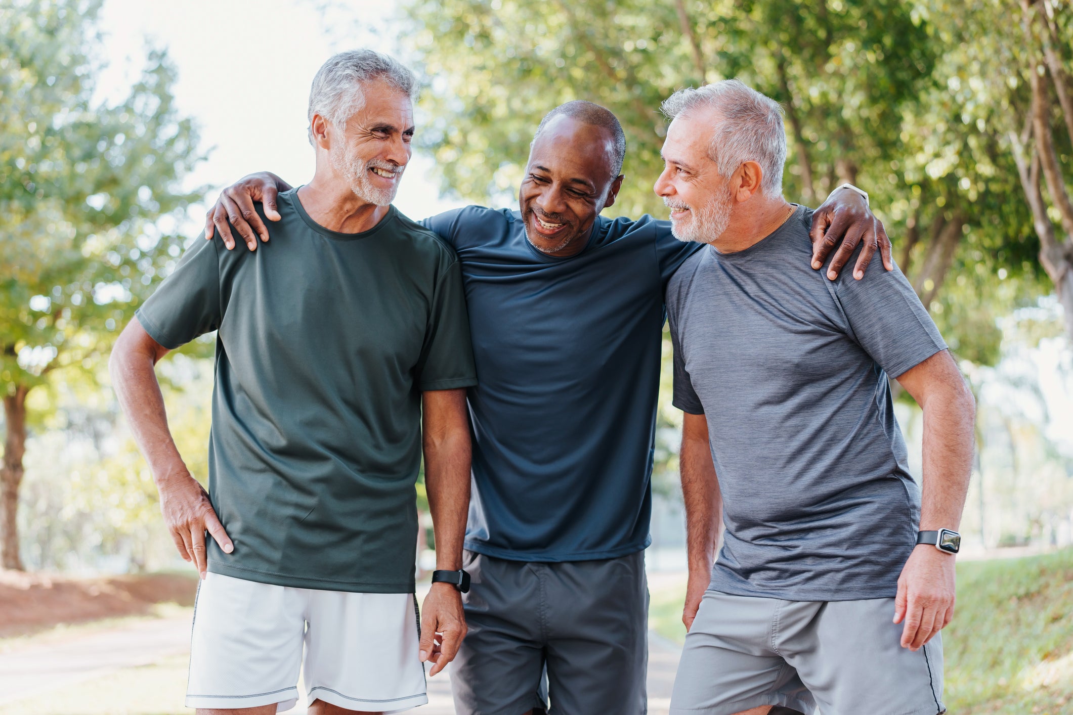 Three middle-aged men standing together outdoors in a park, smiling and laughing with their arms around each other. They are wearing athletic t-shirts in dark green, navy blue, and heather gray, paired with gray shorts.