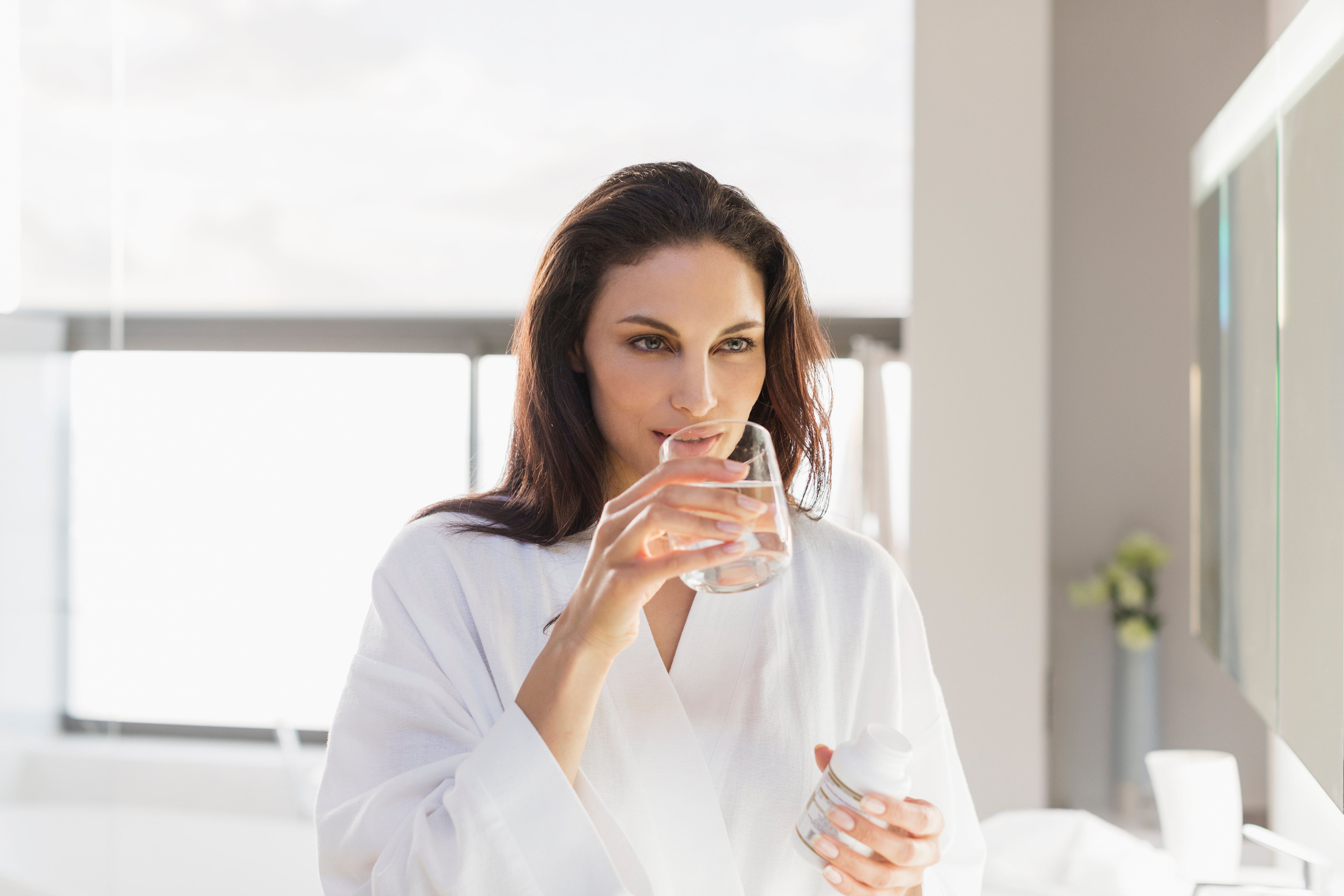 Woman drinking a glass of water in a bright bathroom during her morning skincare and wellness routine.