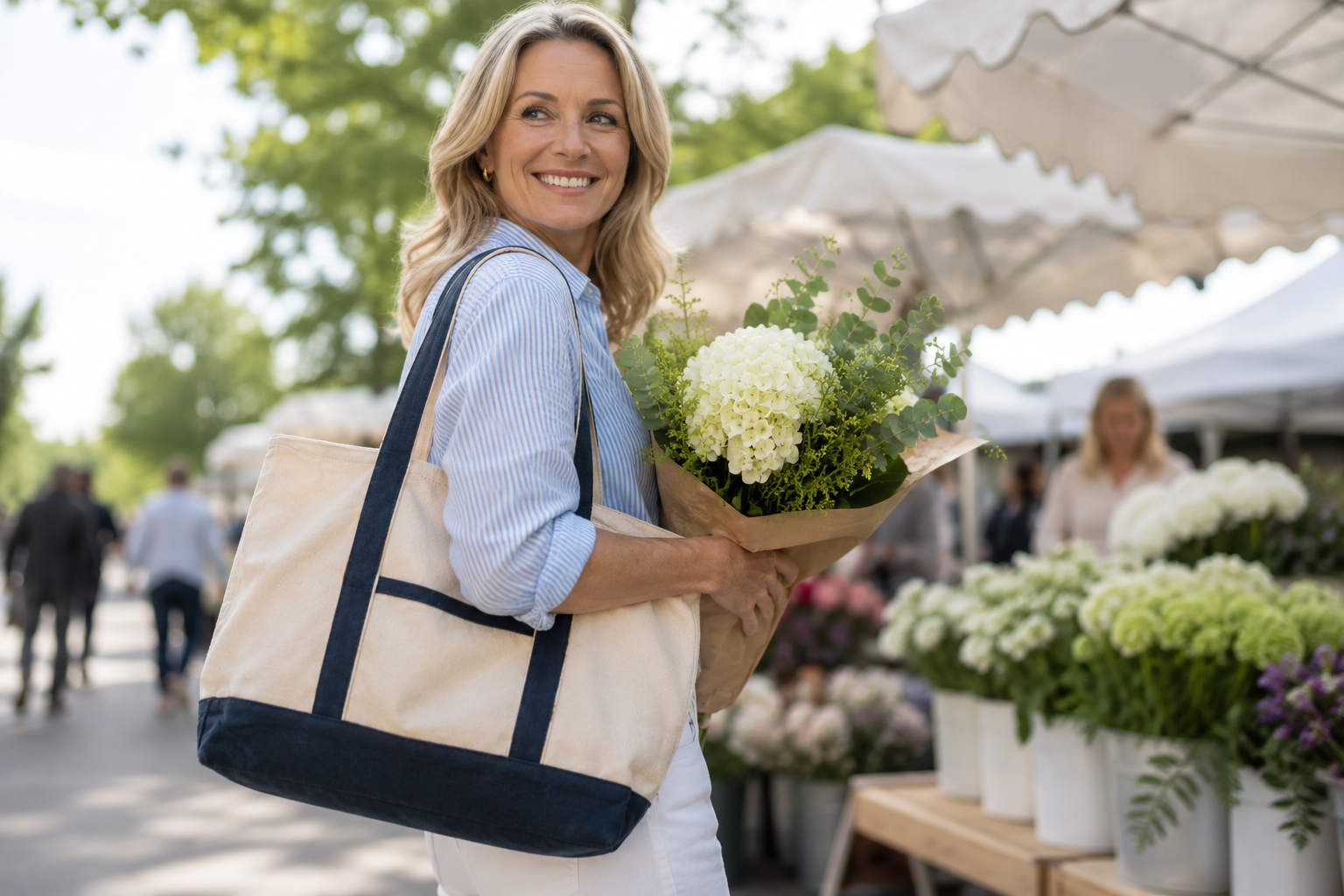 A smiling woman at an outdoor flower market carries a large cream canvas tote bag with dark navy blue trim and handles, while holding a bouquet of white flowers.