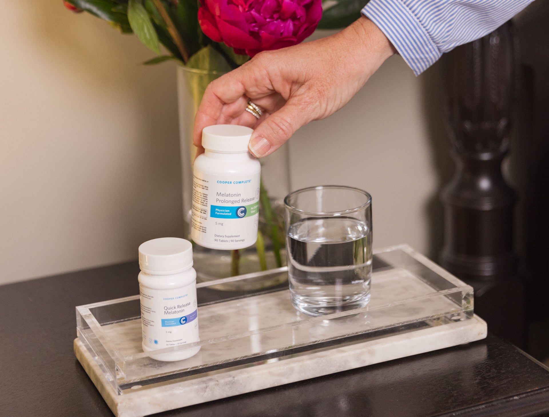 Hand reaching for a bottle of Cooper Complete Prolonged Release Melatonin beside a glass of water, with Cooper Complete Quick Release Melatonin in the foreground.