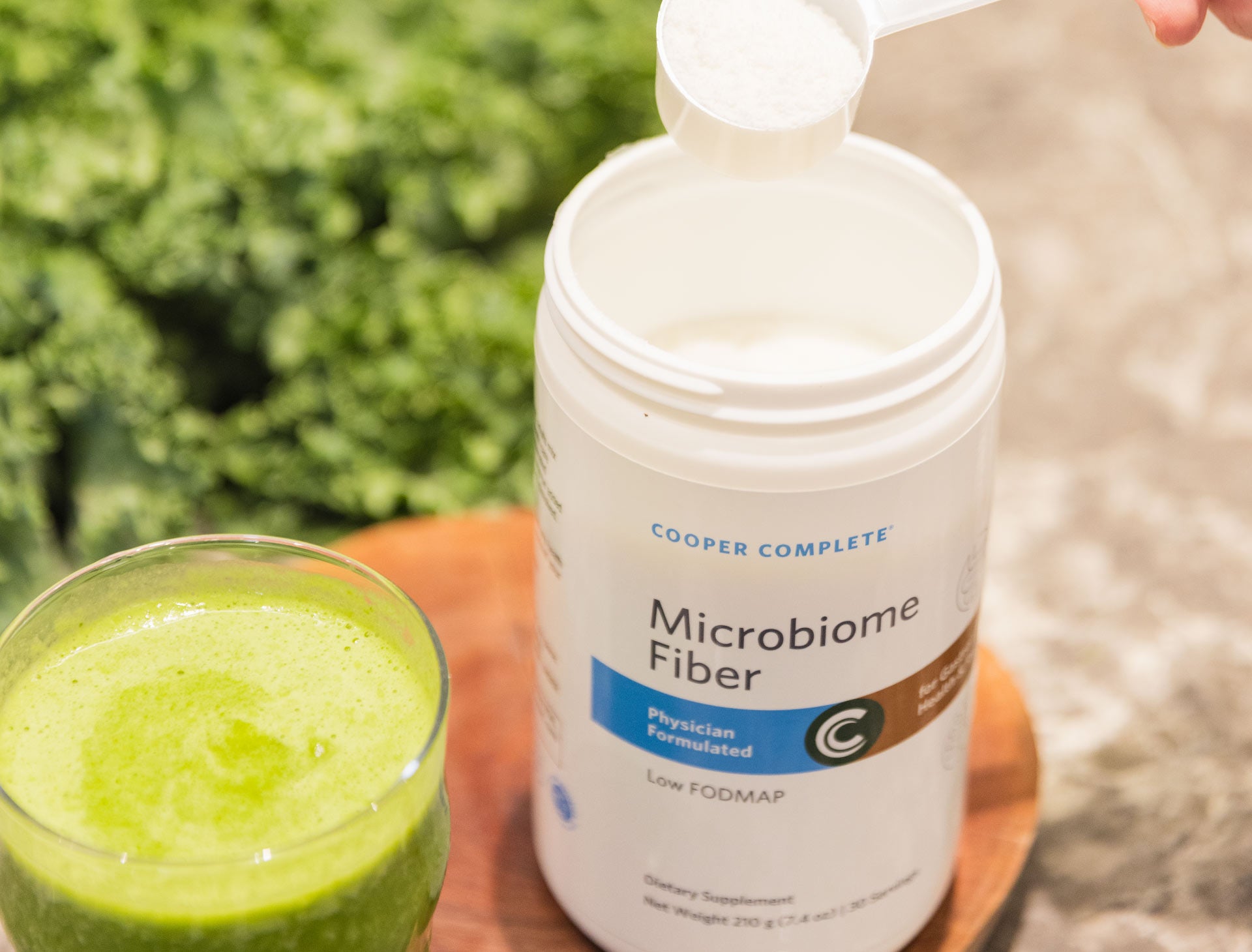 Canister of Cooper Complete Microbiome Fiber supplement next to a glass of green juice with vegetables in the background.