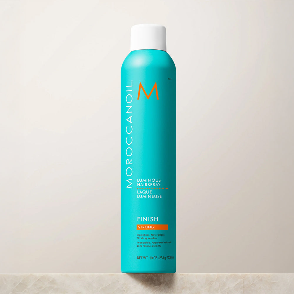 A tall turquoise Moroccanoil Luminous Hairspray can with a white cap, standing upright against a light beige wall on a pale stone surface.