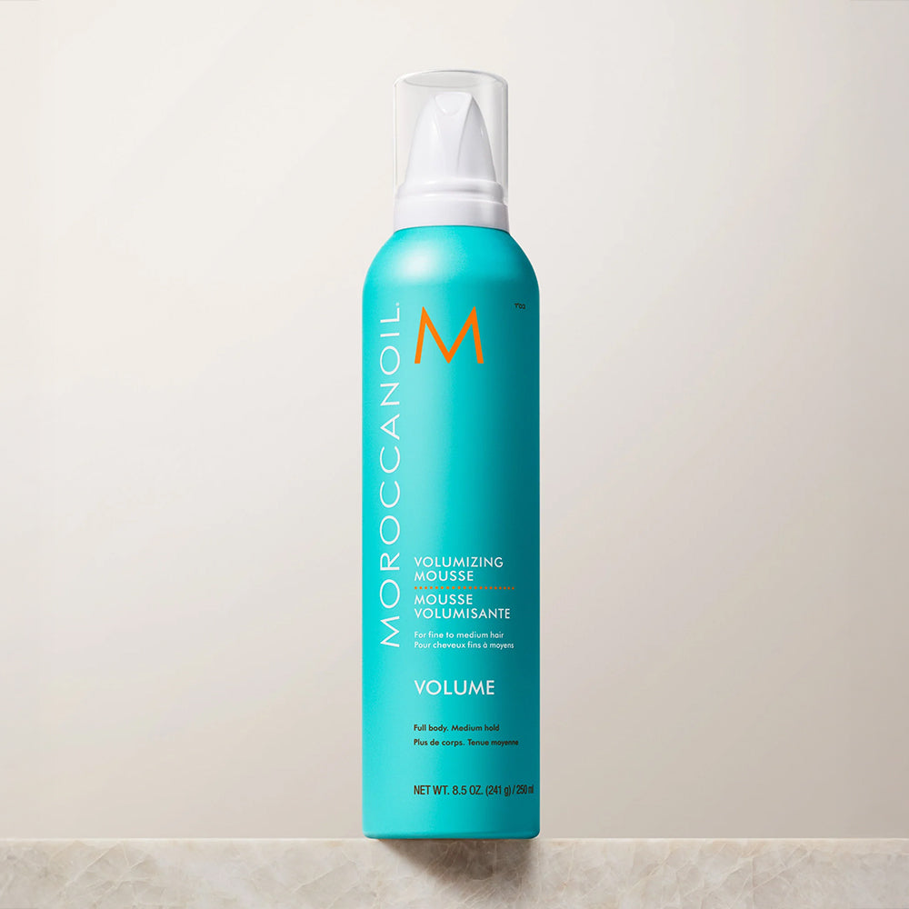 A turquoise Moroccanoil Volumizing Mousse can with a white pump cap, standing upright on a light marble surface against a neutral beige background.