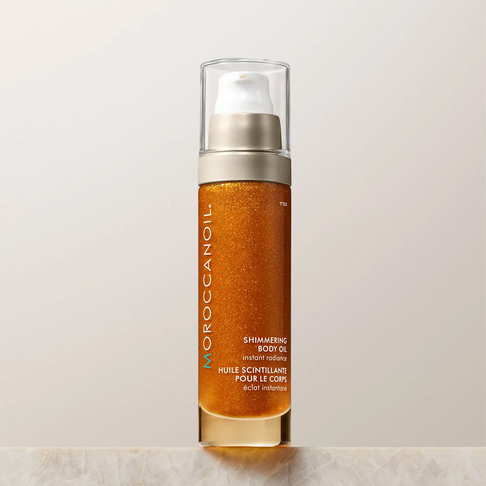 Moroccanoil Shimmering Body Oil in a clear rectangular bottle with a gold glitter liquid inside, a metallic gold pump, and a clear cap, set against a light beige background.