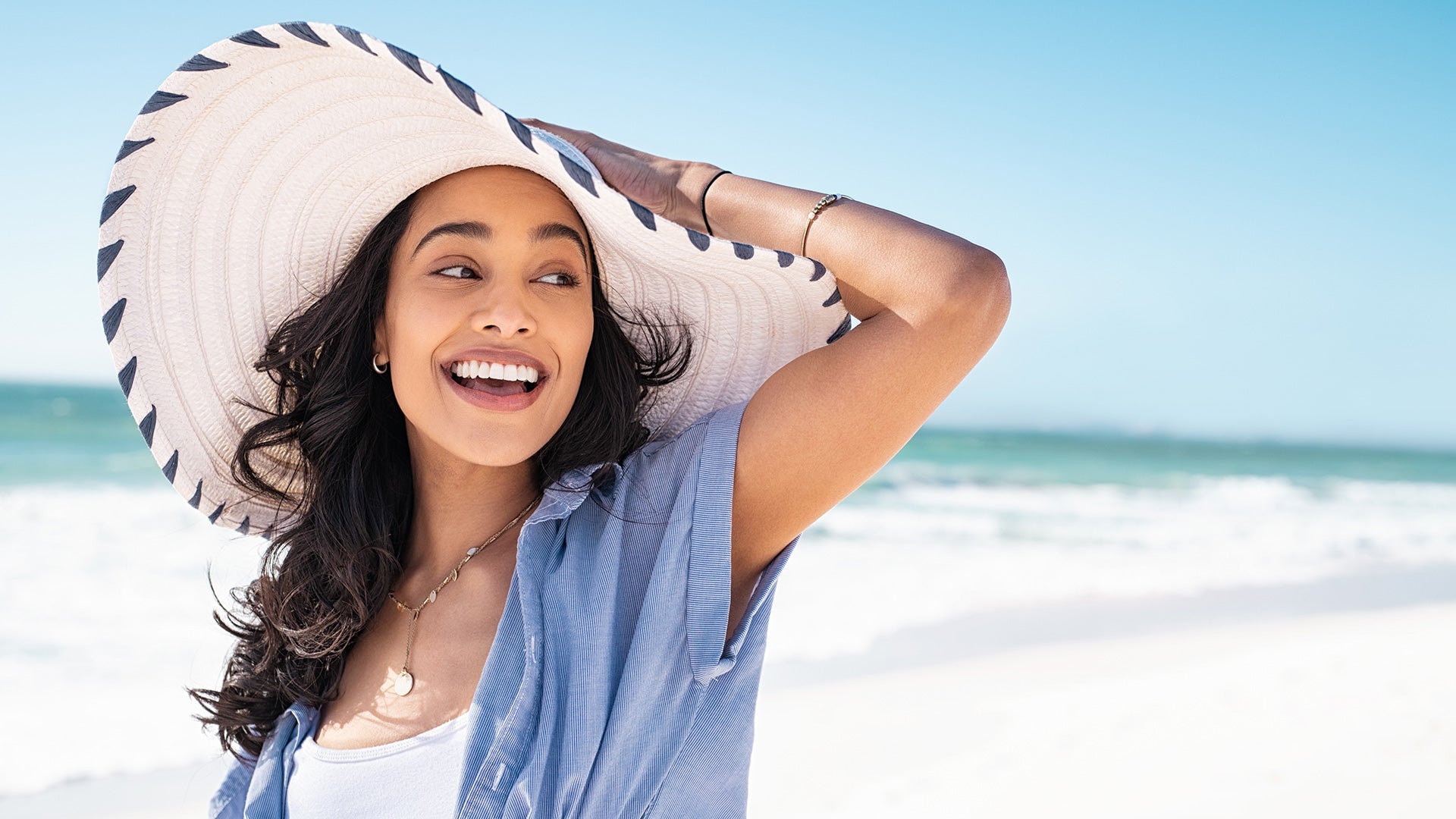 Woman wearing a wide-brimmed sun hat while smiling on the beach on a sunny day.