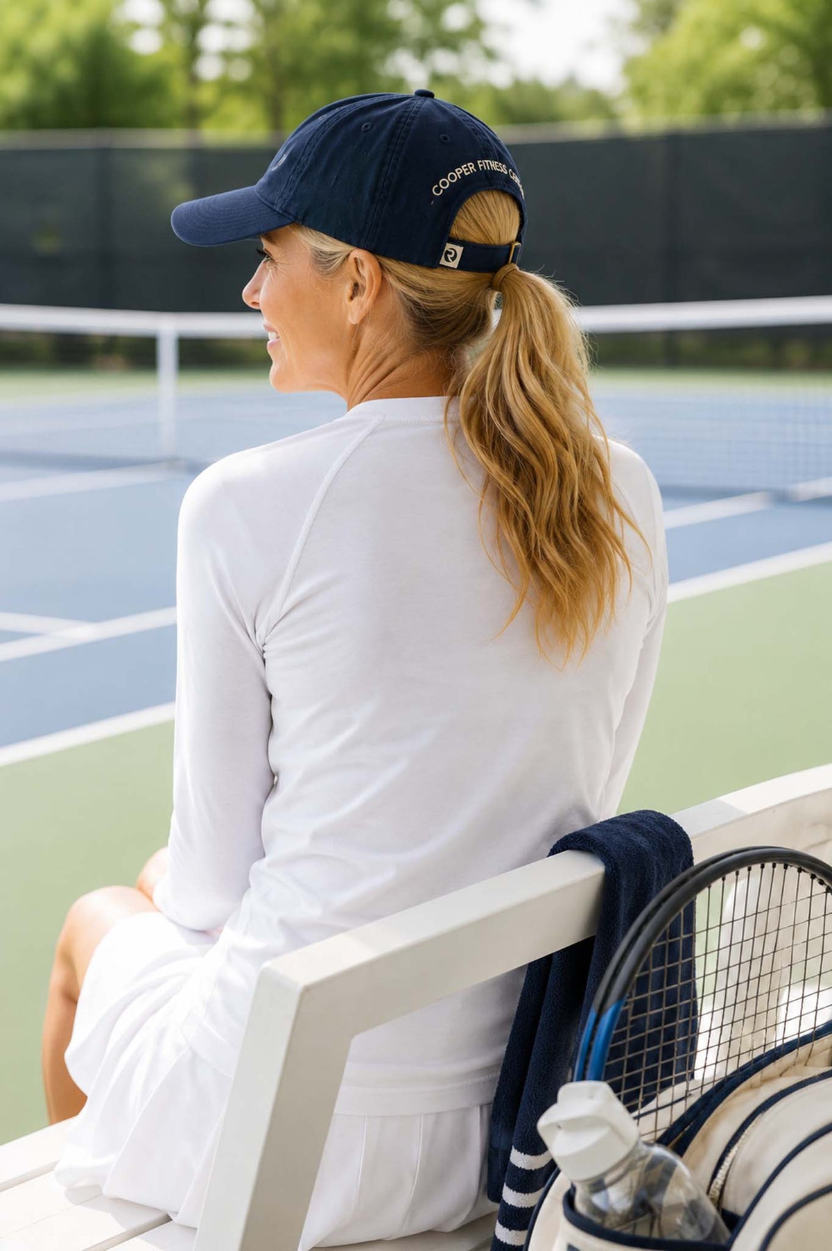 A woman sitting on a white tennis court bench, seen from behind, wearing a white tennis outfit with a navy ball cap that has "Cooper Fitness Center" embroidered on the back. A tennis racket is visible nearby.