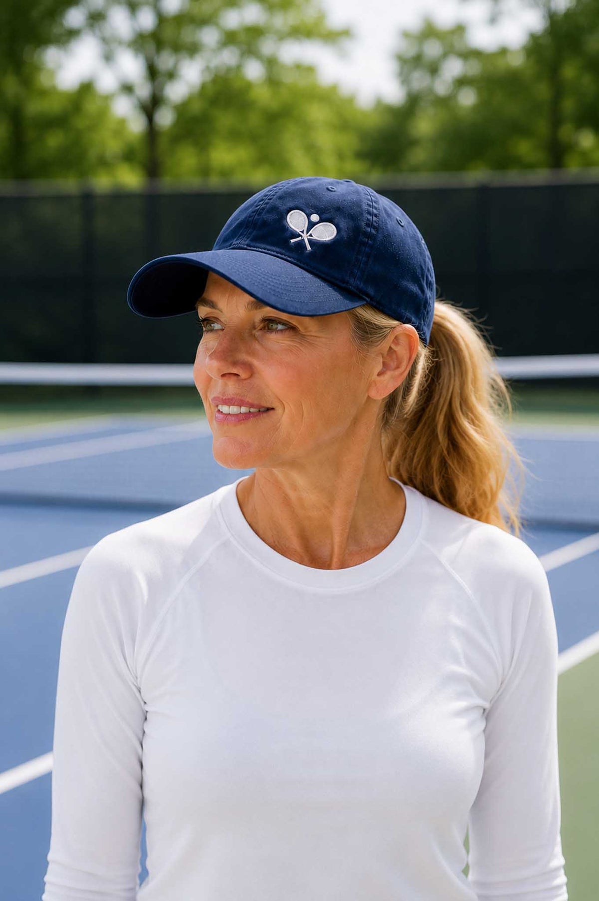 A woman on a tennis court wearing a navy blue baseball cap that has white intersecting tennis rackets and a ball, and a white long-sleeve athletic shirt.