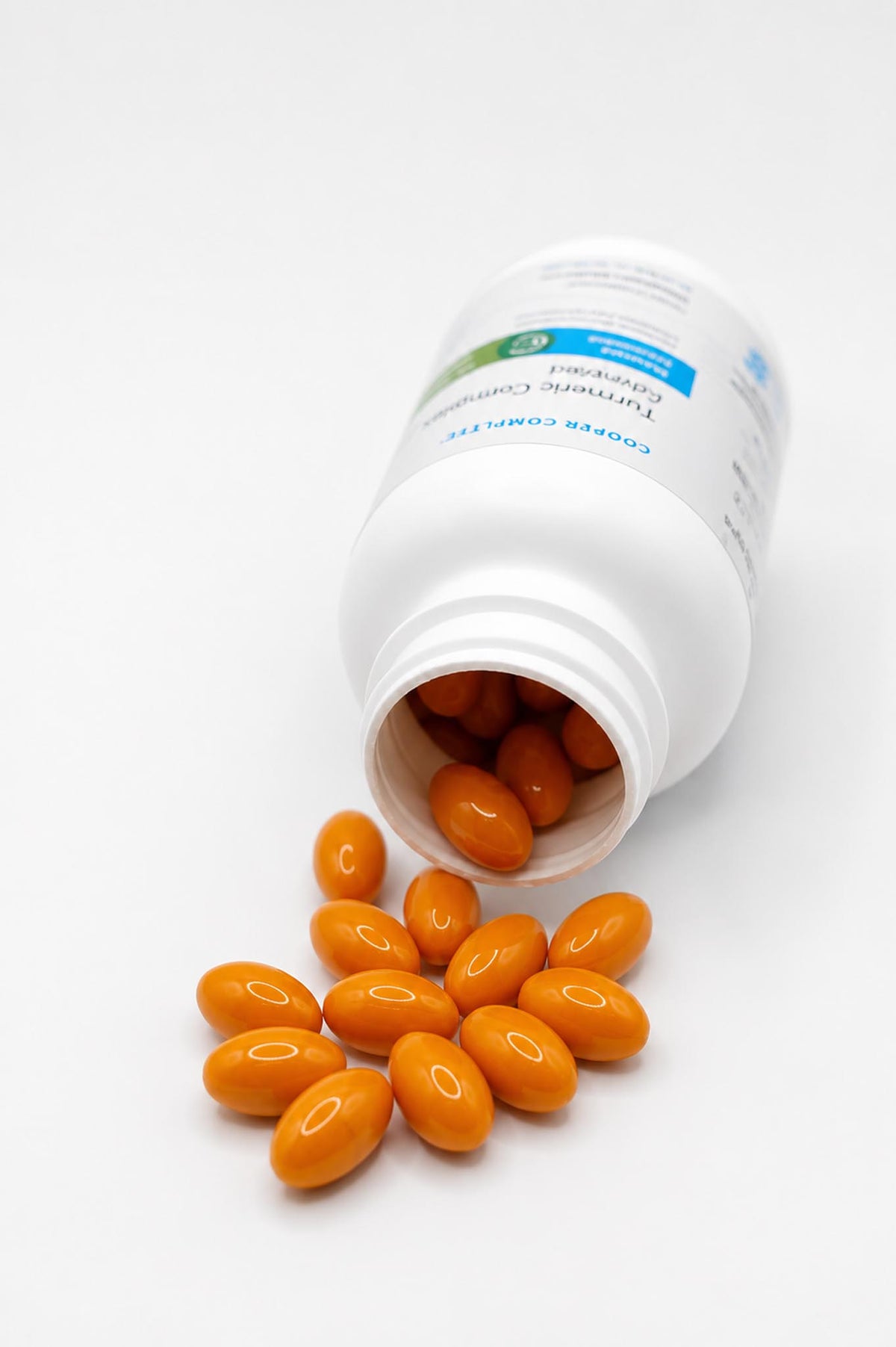 An open white Cooper Complete Turmeric Curcumin Complex supplement bottle lying on its side with several orange oval capsules spilling out onto a white surface.