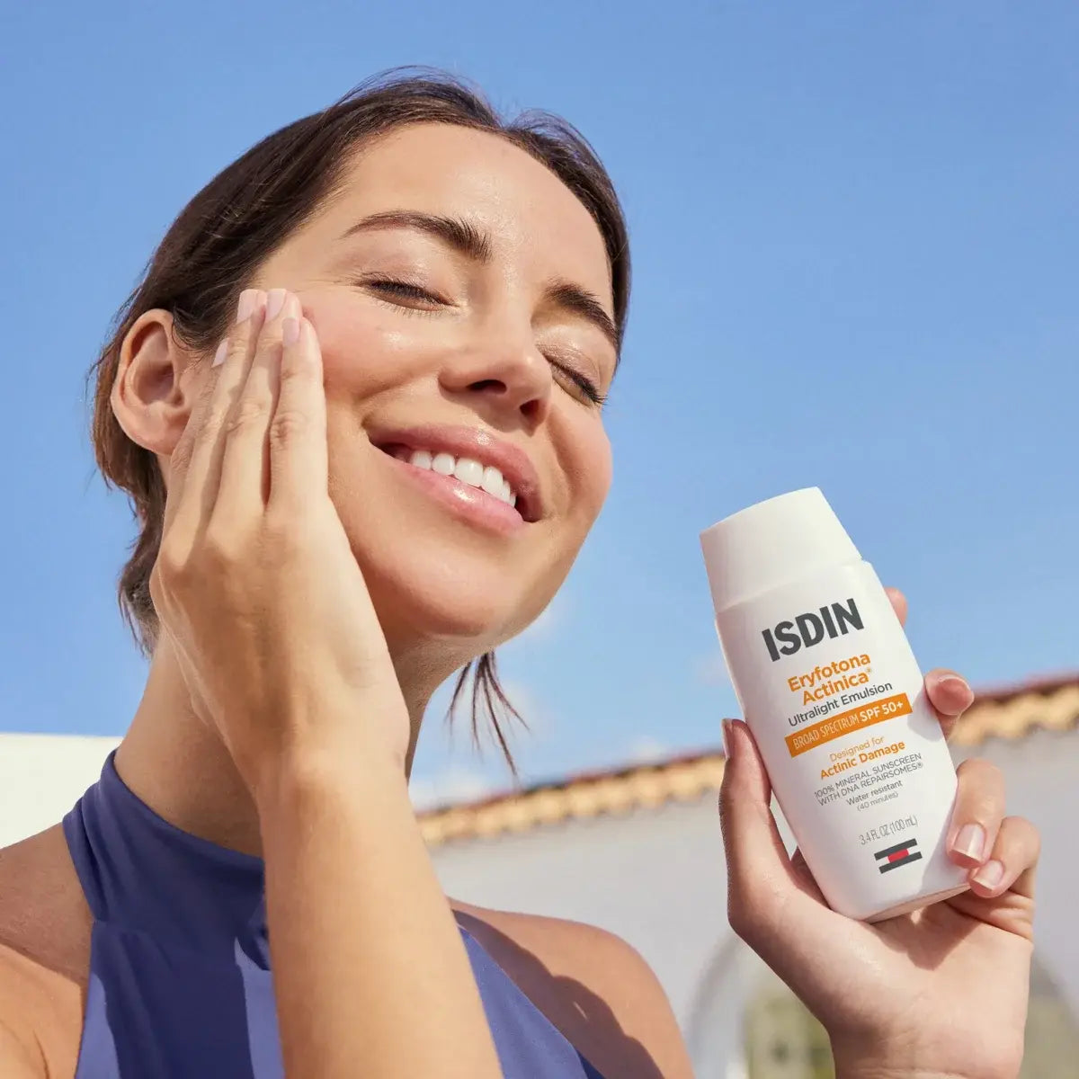 Smiling brunette woman rubs ISDIN Eryfotona Actinica ultralight emulsion SPF 50+ onto face in front of a bright blue sky.