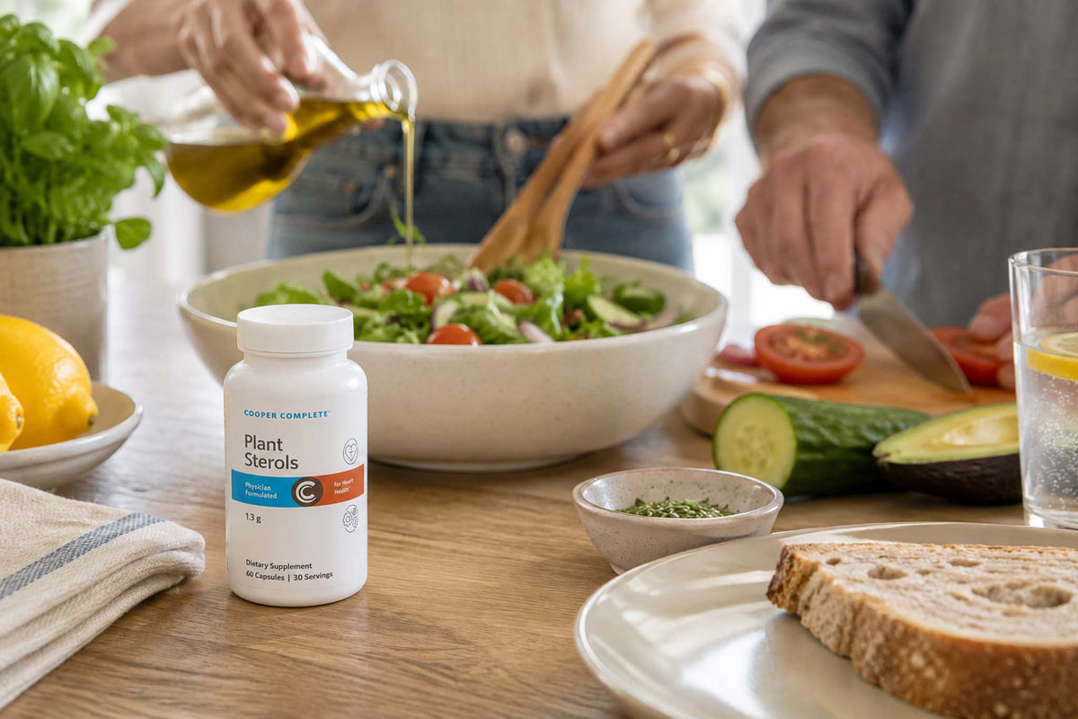 A bottle of Cooper Complete Plant Sterols supplement bottle on a kitchen counter beside a fresh salad as two adults prepare a healthy meal.