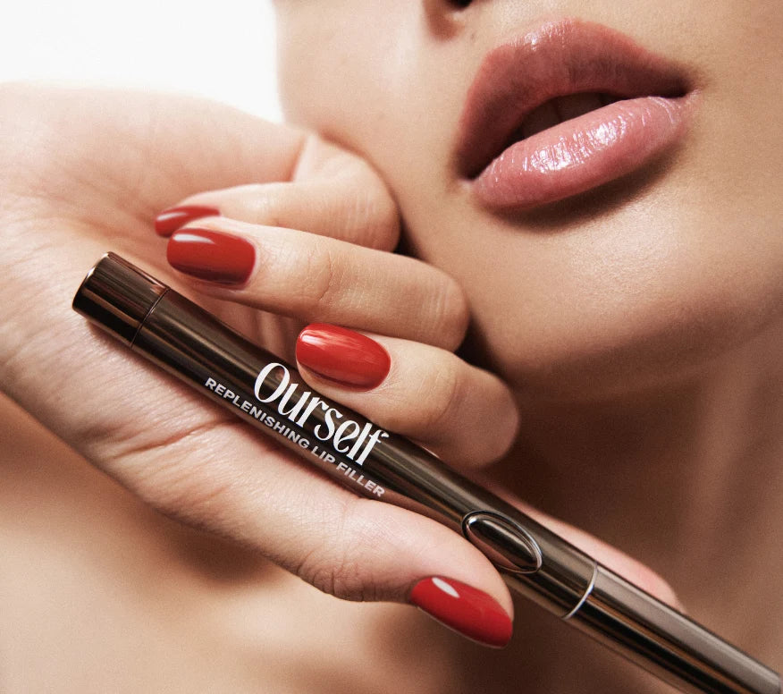 Close up of woman with red nails holding Ourself Replenishing Lip Filler against her face with healthy-looking lips.