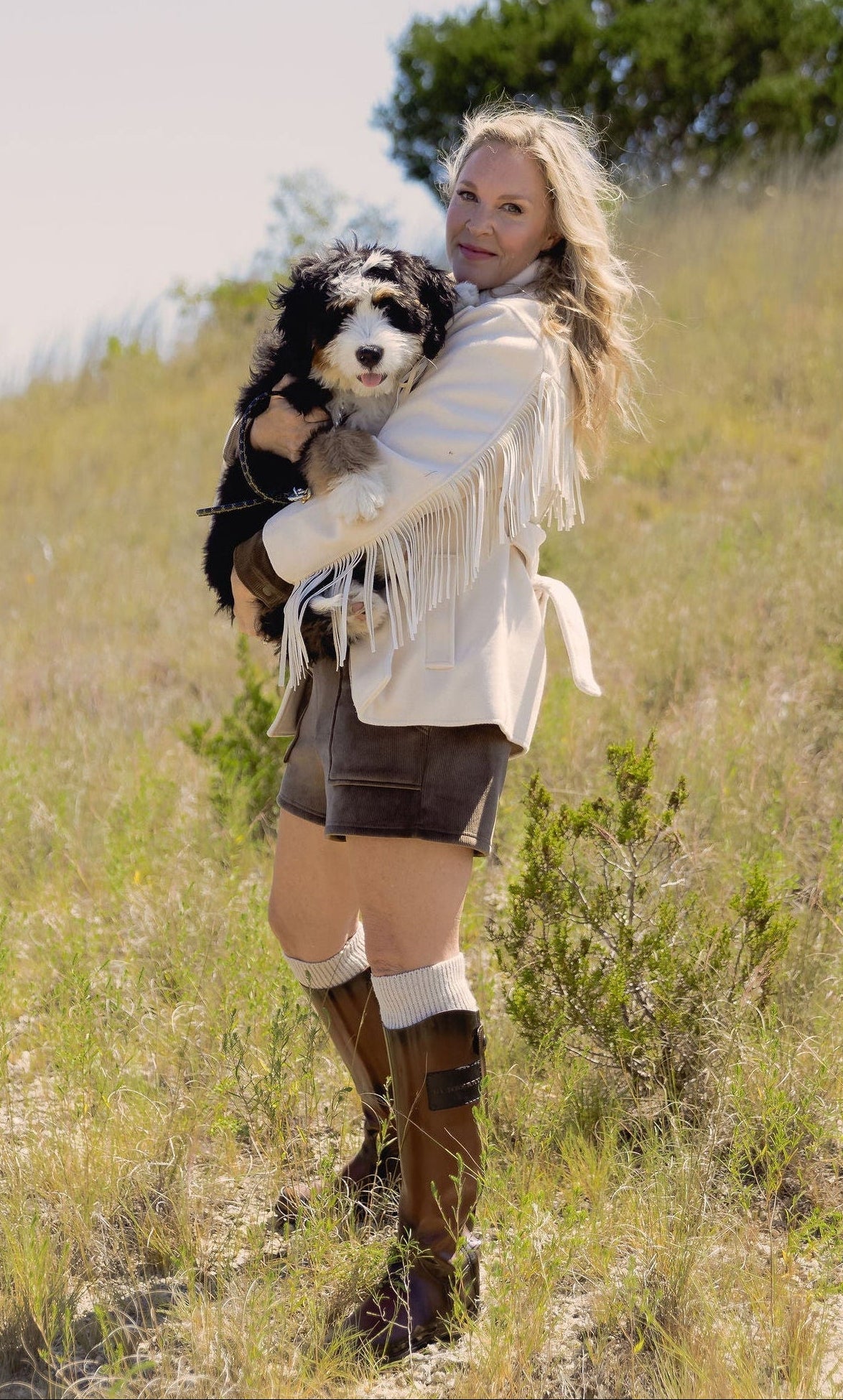 Model wearing the BLANK NYC Rodeo Star Blazer in Off White with Be Fit brown shorts, and tall brown boots while holding a dog in a grassy field.