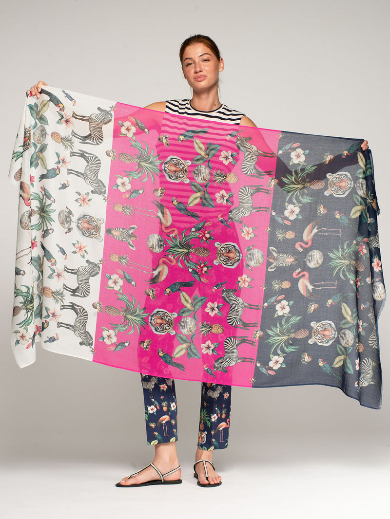 Model holding Vilagallo Jungle Disco oversized lightweight semi-sheer cotton-modal scarf featuring a vibrant tropical jungle print with colorful birds, exotic animals, lush foliage, and bold graphic stripes.