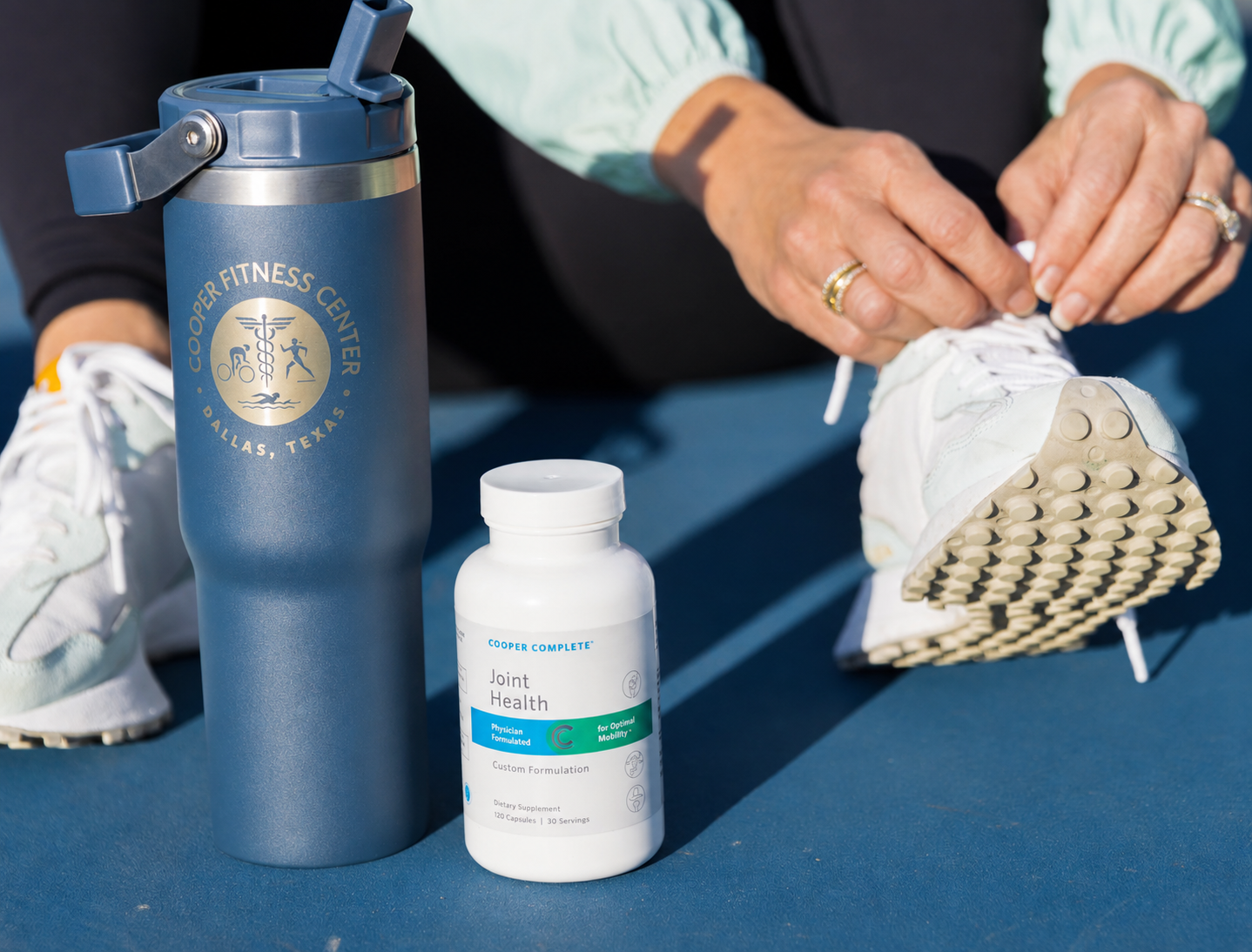 A woman outdoors on a blue court, tying the laces of a white sneaker while a large navy insulated Cooper Fitness Center bottle and a bottle of Cooper Complete Joint Health supplement sit in the foreground.