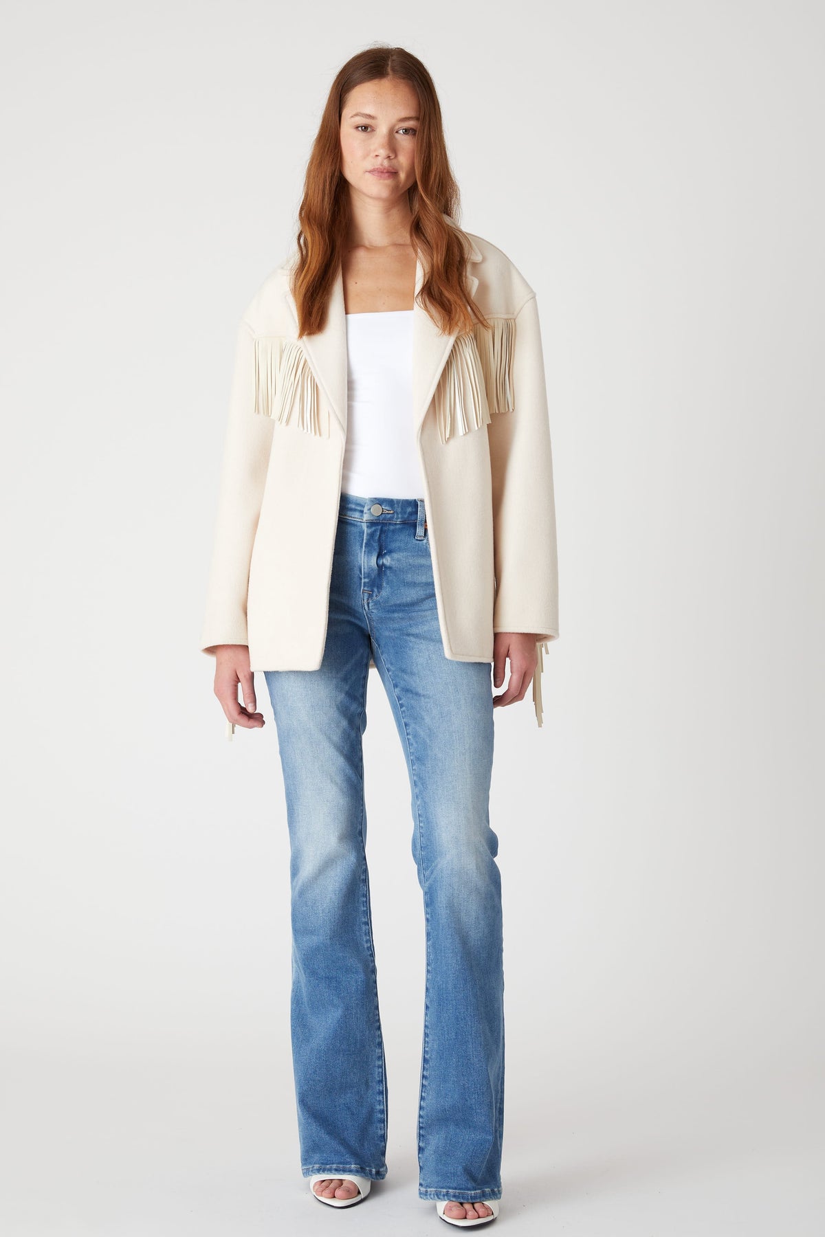 Model wearing the BLANK NYC Rodeo Star Blazer in Off White open over a white top, blue jeans and sandals.