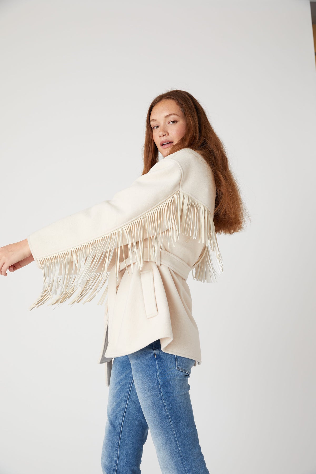 Side view of a model wearing the BLANK NYC Rodeo Star Blazer in Off White with one arm extended to show sleeve fringe.