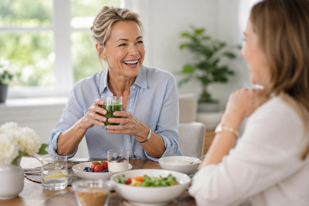 A smiling woman in a light blue button-up shirt holds up a glass of green juice while sitting at a dining table with bowls of fruit and vegetables, conversing with another woman in a bright, modern kitchen setting.