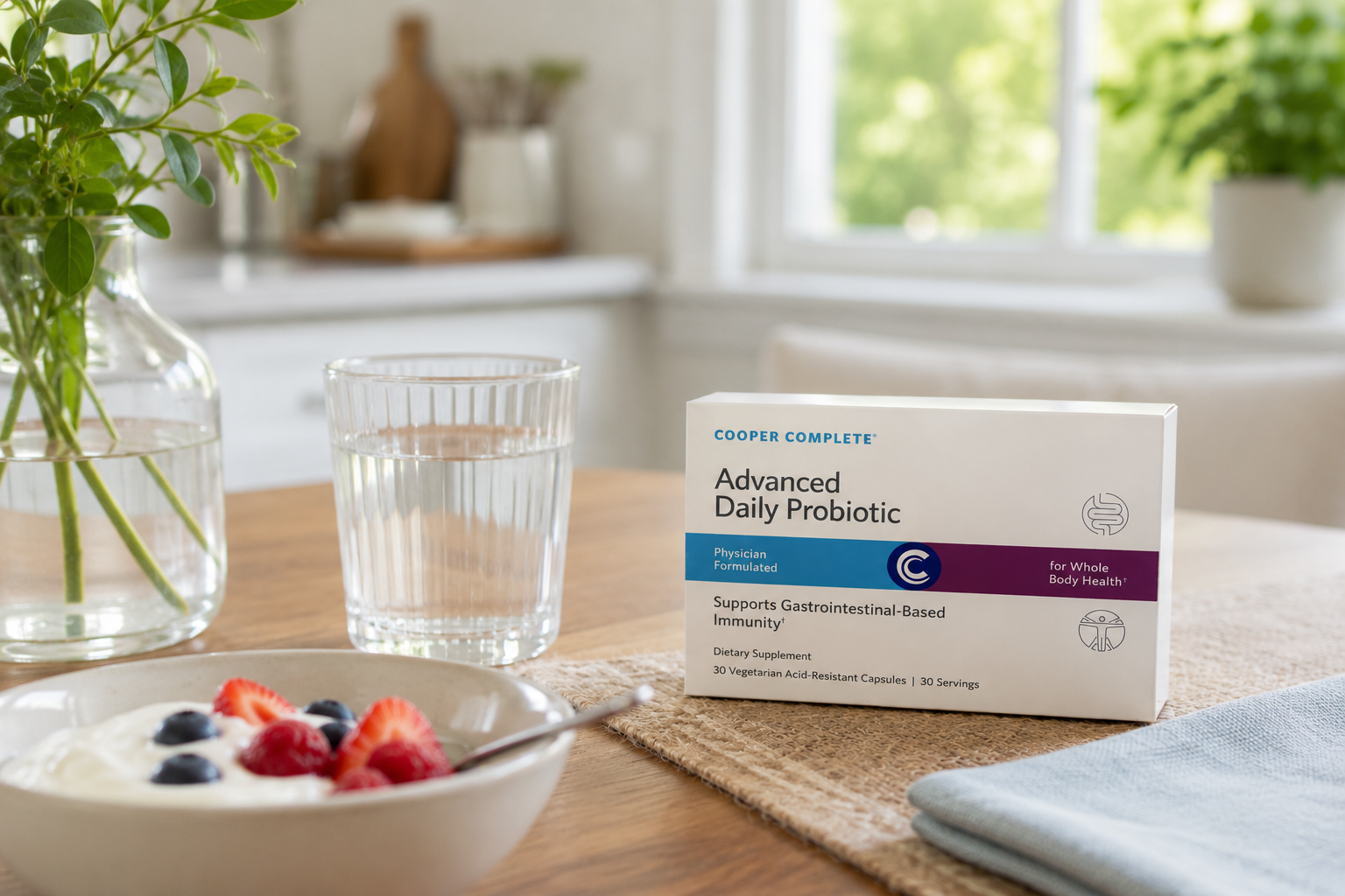 A box of Cooper Complete Advanced Daily Probiotic capsules sits on a wooden table next to a glass of water, a bowl of yogurt with berries, and a vase with greenery in a bright kitchen setting.