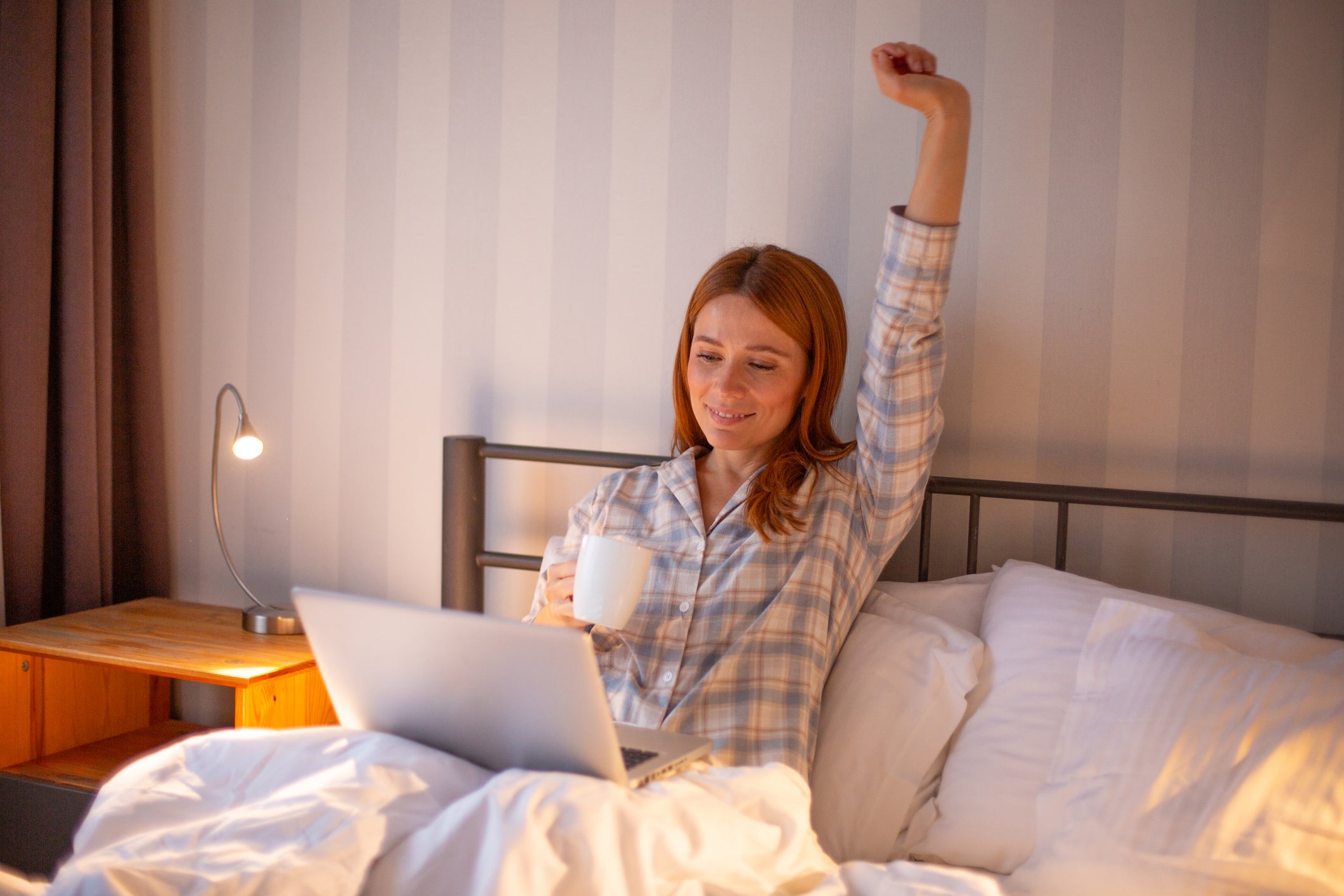 A woman in a plaid pajama shirt sits in bed with white bedding, holding a white mug and using a laptop, with one arm raised in the air. A bedside lamp is turned on next to her.