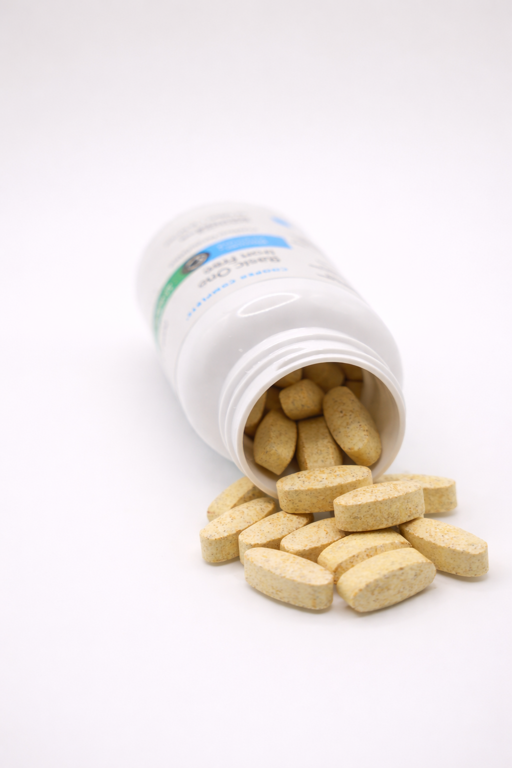 A white plastic Cooper Complete Basic One Iron Free daily multivitamin bottle lying on its side with light tan oval tablets spilling out onto a white surface.