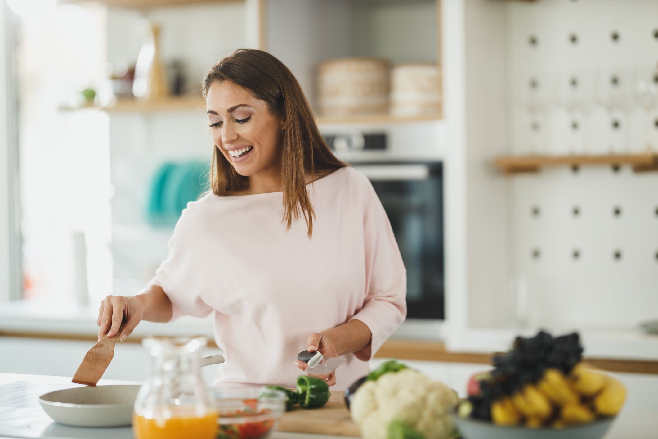 A woman with long brown hair, wearing a light pink top, is smiling while cooking in a bright, modern kitchen. She is standing behind a counter with various fresh ingredients, including a jar of orange juice, a green pepper, and a head of cauliflower.