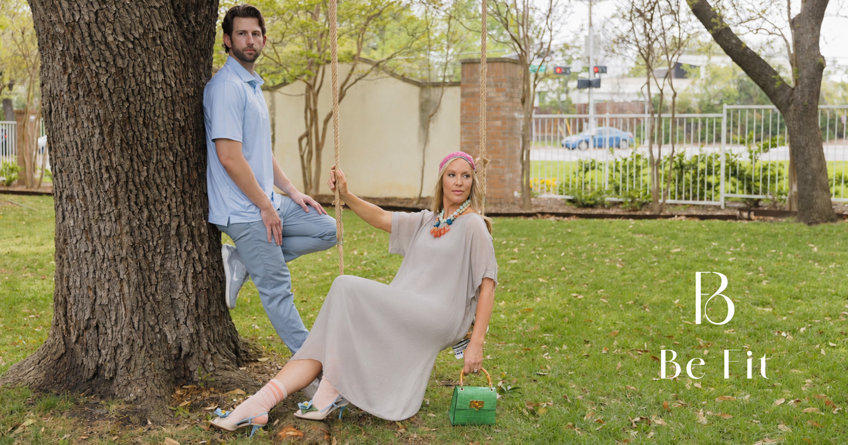 Man and woman modeling Be Fit™ apparel and accessories in a park, with the woman seated on a rope swing and the man leaning against a tree.