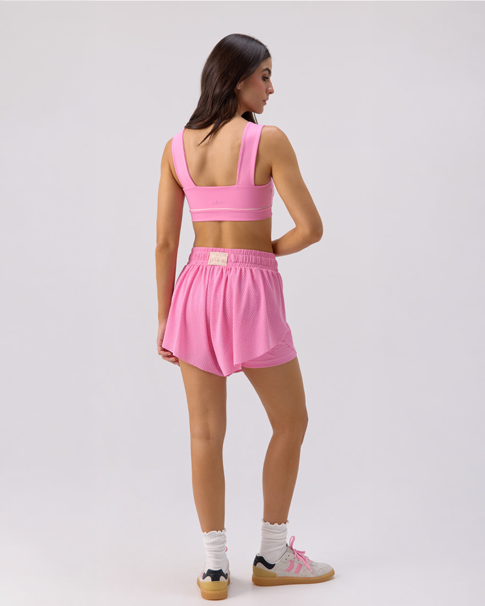 Back view of a model wearing a Glee x Be Fit pink athletic outfit of sports bra and high-waisted mesh shorts on a white background