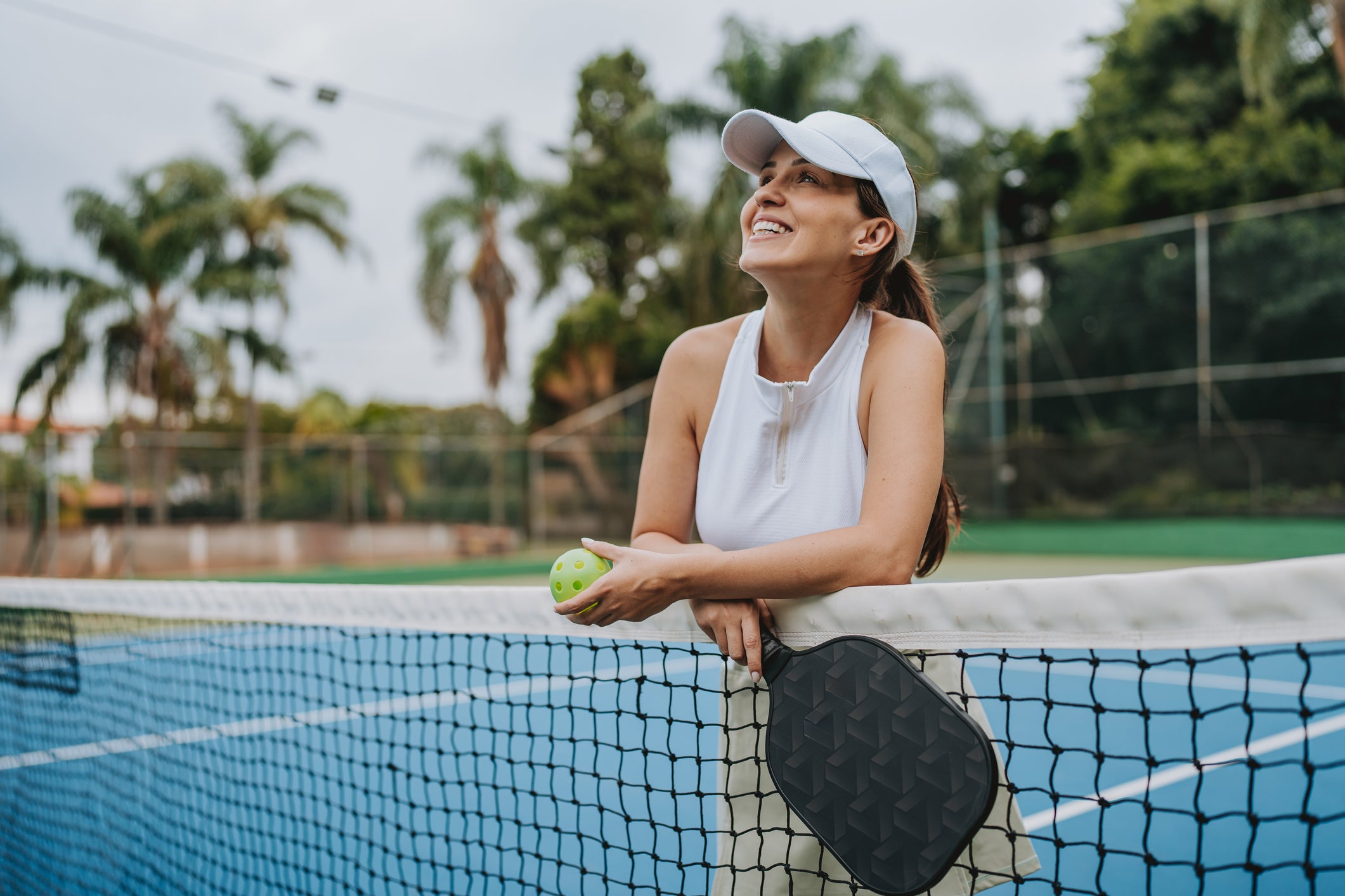 A woman on a pickleball court wearing a white sleeveless top and white cap, holding a pickleball paddle and ball, with palm trees in the background.
