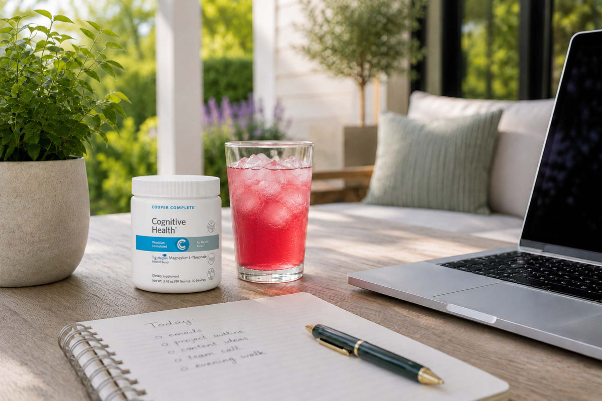 A canister of Cooper Complete Cognitive Health supplement sits on a wooden table next to a glass of pink iced drink, a notebook with a pen, and an open laptop in a bright, modern home outdoor setting.