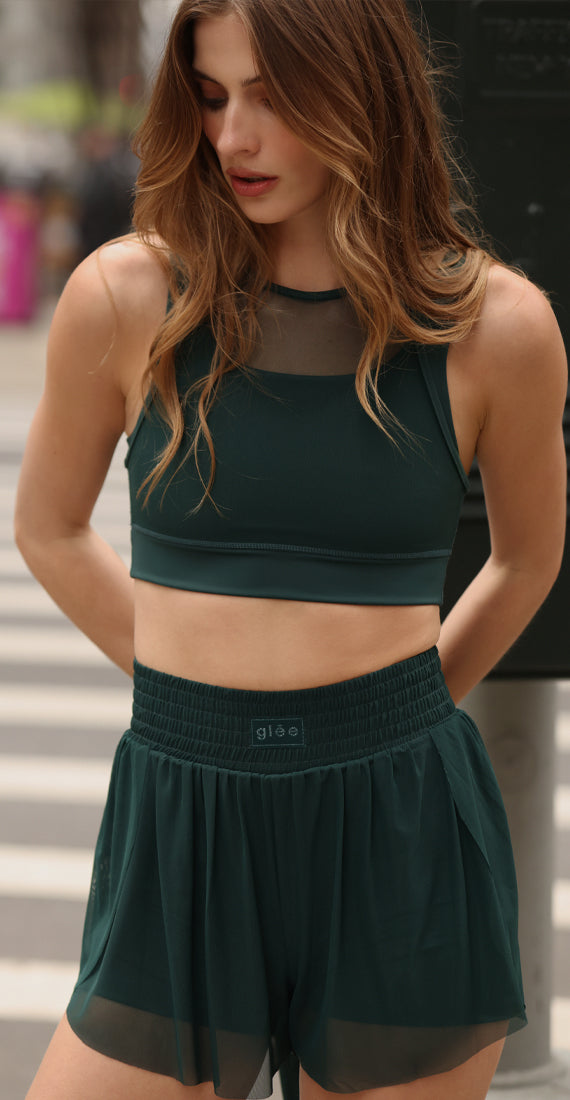 Model wearing the Be Fit x Glee botanic green Ballerina Mesh Shorts with a matching mesh-panel Siren Lift Top sports bra outdoors.
