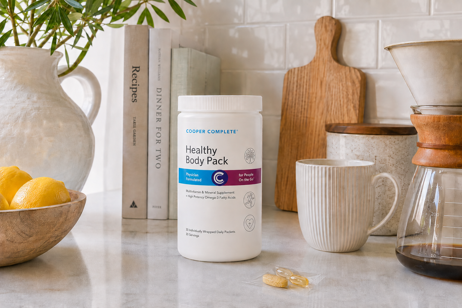 A white tub of Cooper Complete Healthy Body Pack supplement stands on a kitchen counter, surrounded by a mug, coffee maker, single-serving supplement packet, books, and lemons in a bright, kitchen setting.