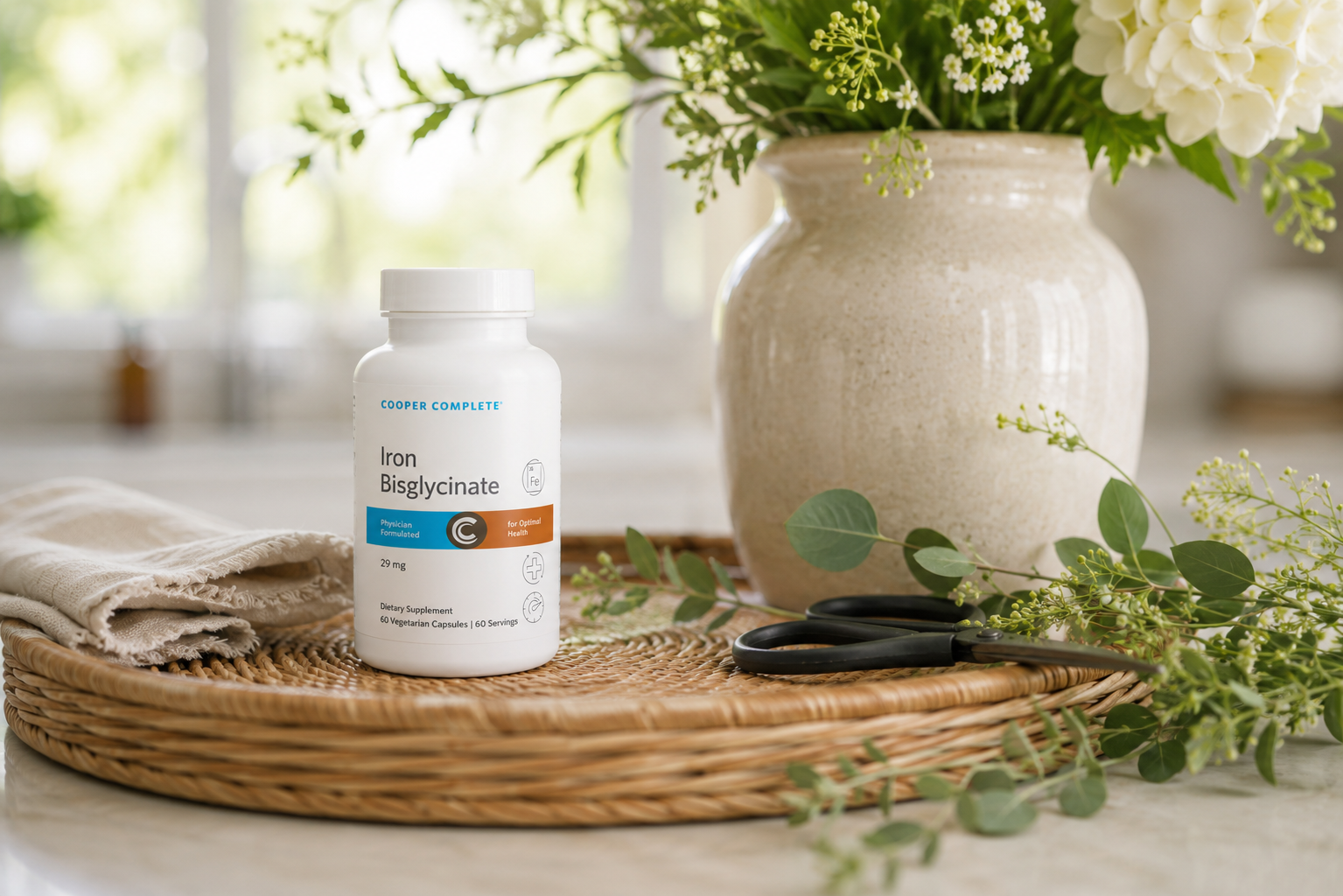 A bottle of Cooper Complete Iron Bisglycinate displays on a woven rattan tray beside a fresh flower arrangement on a bright kitchen countertop. 