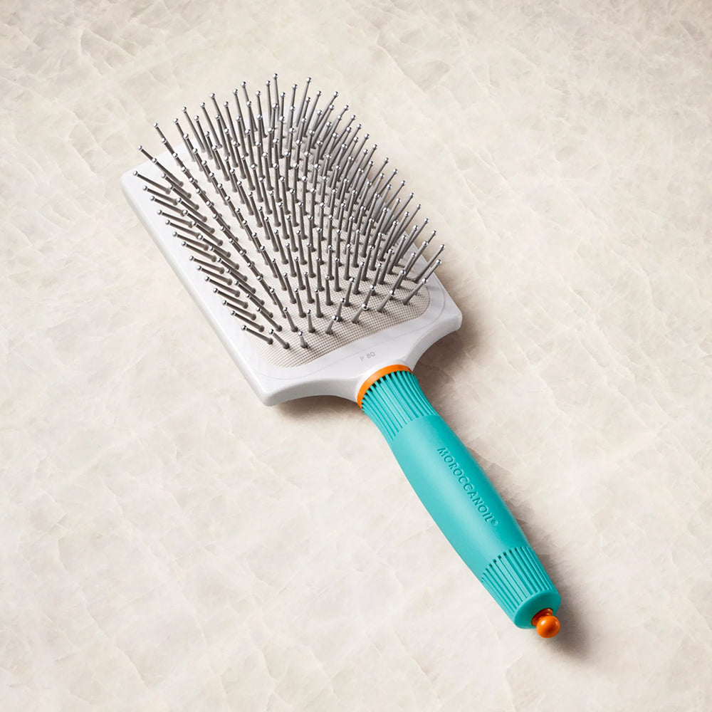 A Moroccanoil paddle hairbrush with a white rectangular head, silver metal bristles, a turquoise handle, and a small orange accent, shown on a light beige textured background.
