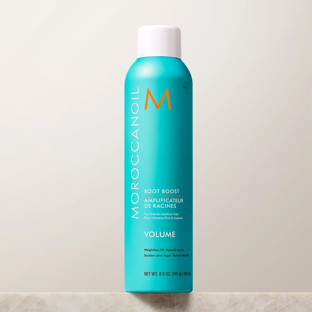 A bottle of Moroccan Oil Volume Root Boost hairspray in a vibrant turquoise color with a white cap, standing on a light marble surface against a neutral beige background.