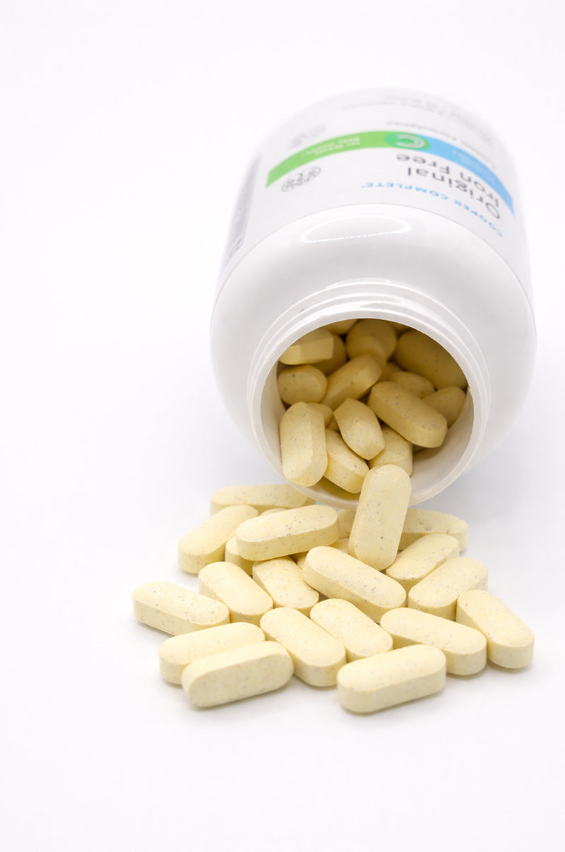 Open white Cooper Complete Original Comprehensive Iron Free Multivitamin pill bottle with tablets spilling out on a white background