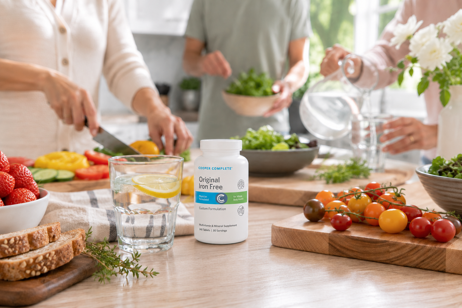 A bottle of Cooper Complete Original Comprehensive Multivitamin Iron Free dietary supplement is displayed on a wooden kitchen table, surrounded by people preparing food in a bright, modern kitchen.
