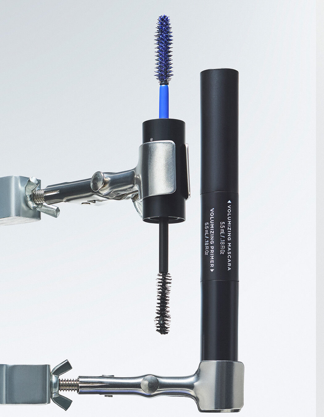 Black tube of RevitaLash Double-Ended Volume Set held by silver metal clamps against studio background with blue Volumizing Primer brush and black Volumizing Mascara brush. 