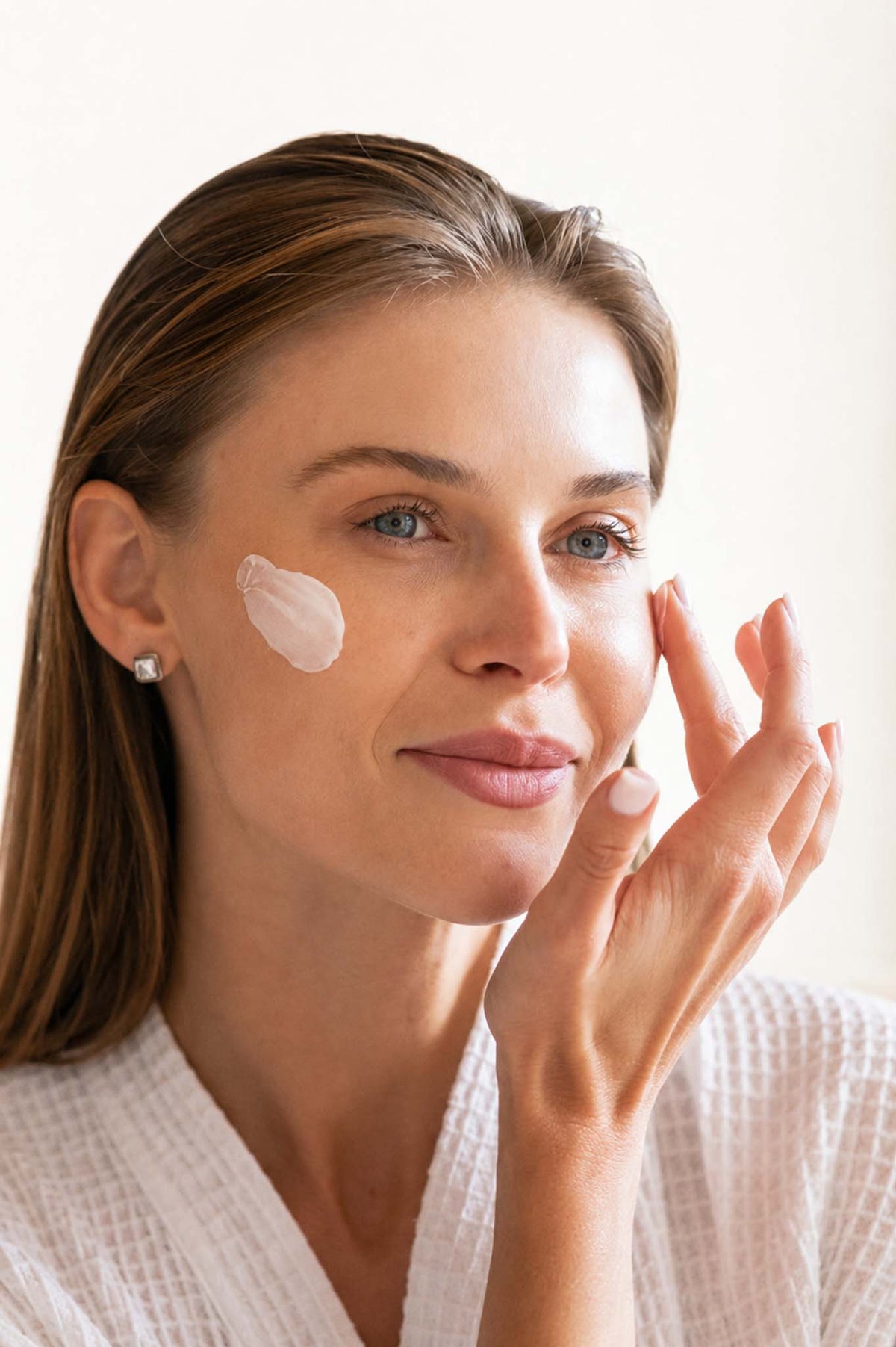 Woman in a white robe applying a lotion or cream to her face, representing skin care.