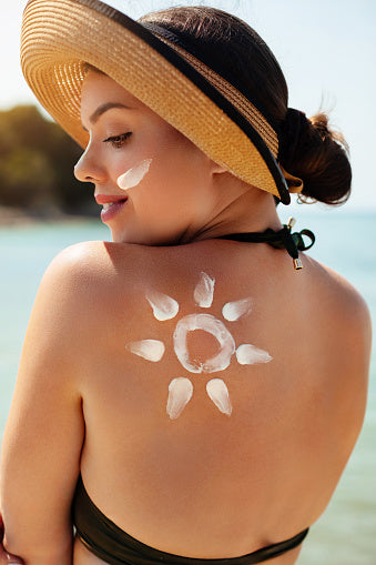 Back view of a woman looking over her shoulder, and there's a stripe of sunscreen painted only her cheek, and onto her back in a stylized sunshine picture.