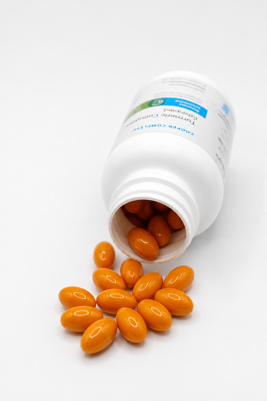 An open white Cooper Complete Turmeric Curcumin Complex supplement bottle lying on its side with several orange oval capsules spilling out onto a white surface.