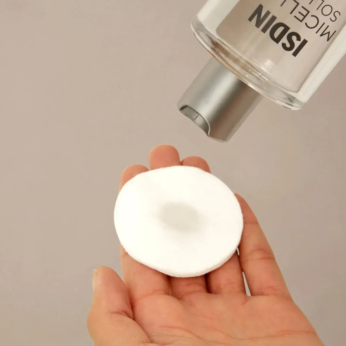 Hand against a beige background holds a cotton pad in palm and apples ISDIN Micellar Solution to the cotton pad. 