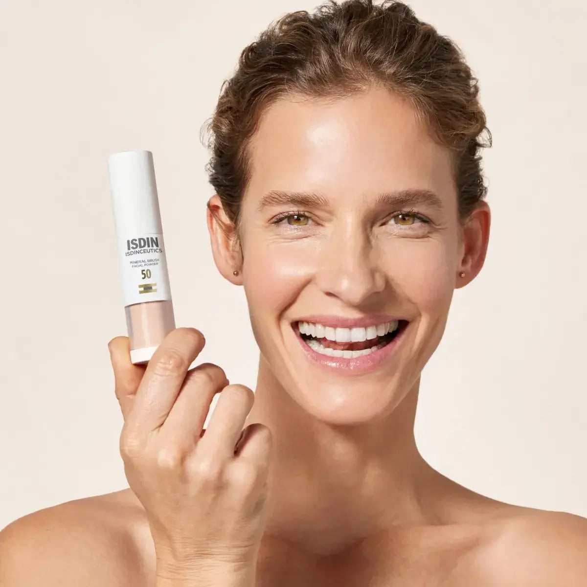 Smiling woman with healthy-looking skin holding a bottle of ISDIN Mineral Brush photoaging defense SPF 50 in front of a light neutral background. 